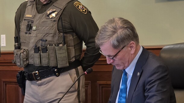 A police officer with his hands behind his back looks down at a desk where the governor is sitting and signing a bill into law.
