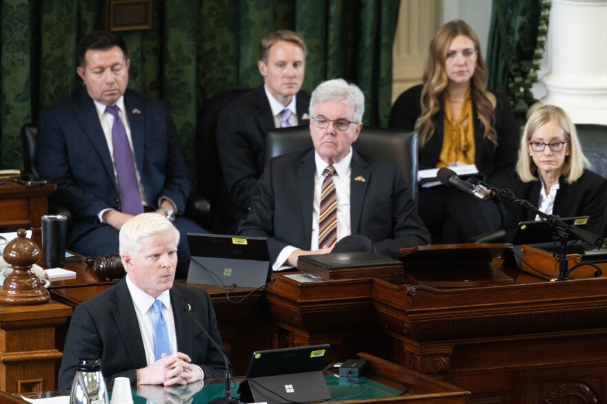 A person in a suit sits at a table with a microphone with people behind him.