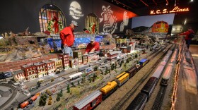 Two men wearing bright red shirts and train conductor caps work amidst a larger display of model trains, tracks and city depictions inside a room decorated for Christmas.