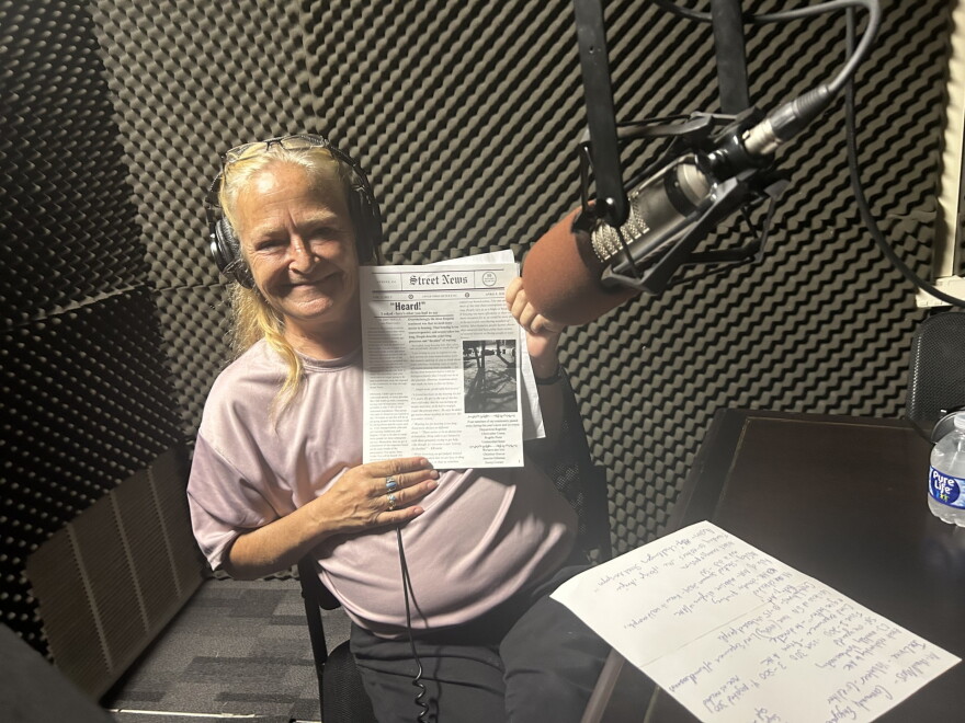 A woman wearing headphones smiles while holding up a copy of Street News.