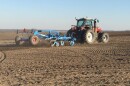 A red tractor hauls a blue, metal-framed planter across a brown, barren field that's criss-crossed with tire marks. 