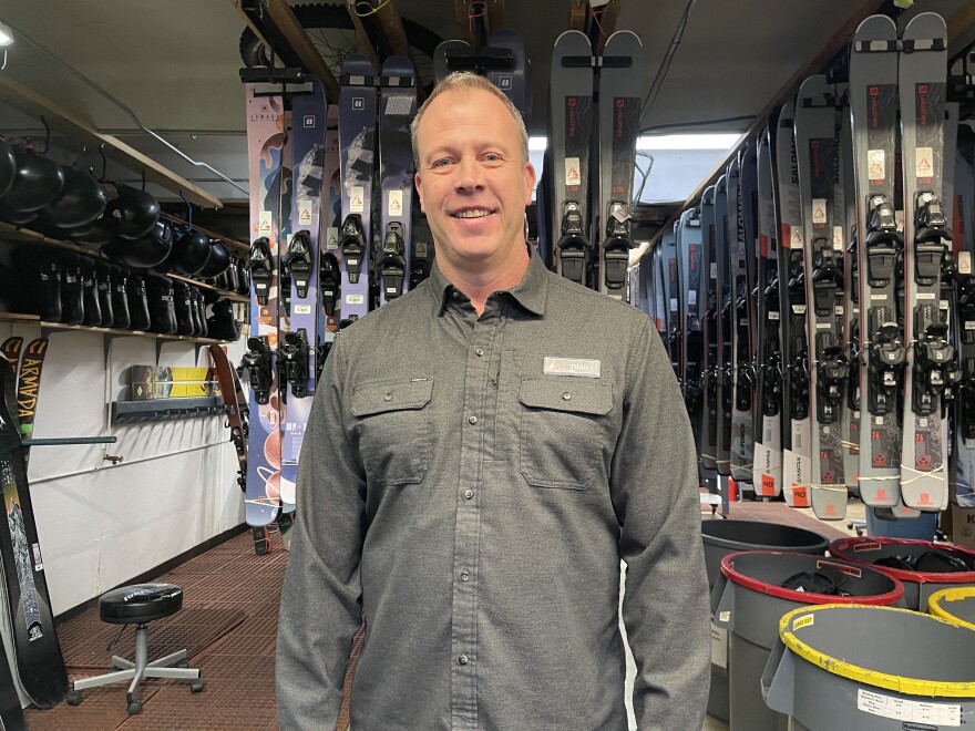 A tall white man with short blonde hair, wearing a long-sleeved gray button down shirt, stands in front of a row of hanging skis. On the right are more rows of skis hanging from the ceiling and big gray trash cans sit underneath. On the left are two shelves, one with black helmets and one with black boots.
