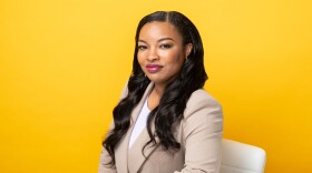 A woman sits at a desk with one arm resting on the table and a bright yellow background behind her.