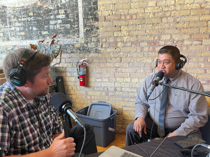 Two men sitting behind microphones and wearing headphones having a conversation. There is a brick wall in the background.