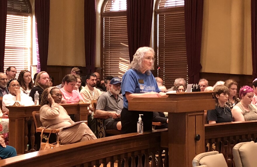 A woman wearing a blue shirt speaks into the microphone on the podium as people fill the rows of seats behind her in the Peoria City Council chambers. 