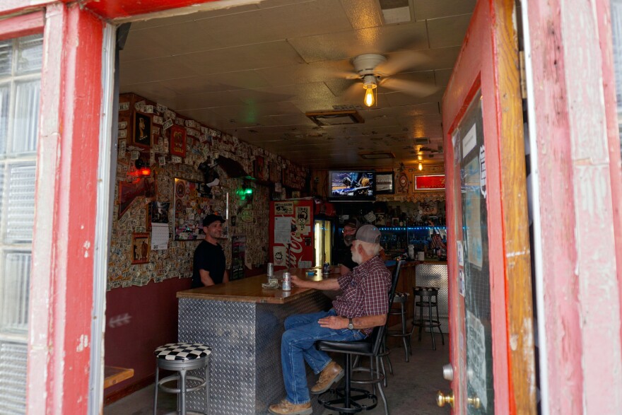 Two people sit inside at a bar. The photo is taken from outside the doorway looking in.