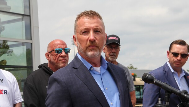 A man in a nice jacket looks serious as he stands behind a microphone. Other men look on. 