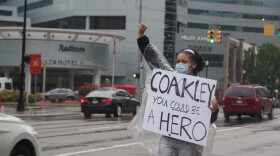 A young woman holds a sign that reads "Coakley, you could be a hero." She is facing a busy street with the gray-clad Radisson Hotel in the background. The street is wet and Custard is wearing a clear plastic poncho.