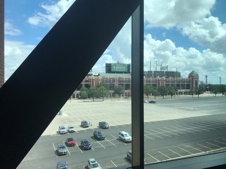 Out of a window, on a sunny day, a large brick stadium can be seen across a parking lot.