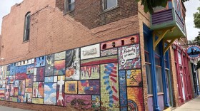 A brown brick building with a colorful mural on one side of it. The mural is a collage of smaller pieces of artwork. The front of the building is blue and purple, with a colorful overhang.