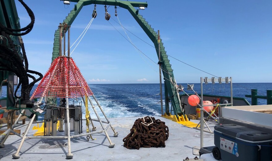 A lot of boat and science equipment on the end of the boat viewing the wake made in deep blue water with a light blue sky behind it. 