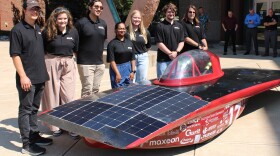 A group of 7 college students stand on a sunny concrete area in front of the solar car