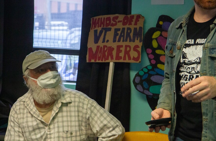 A photo of an older man in a hat and facemask over a white beard, sitting in front of a sign reading hands-off VT. farmworkers.
