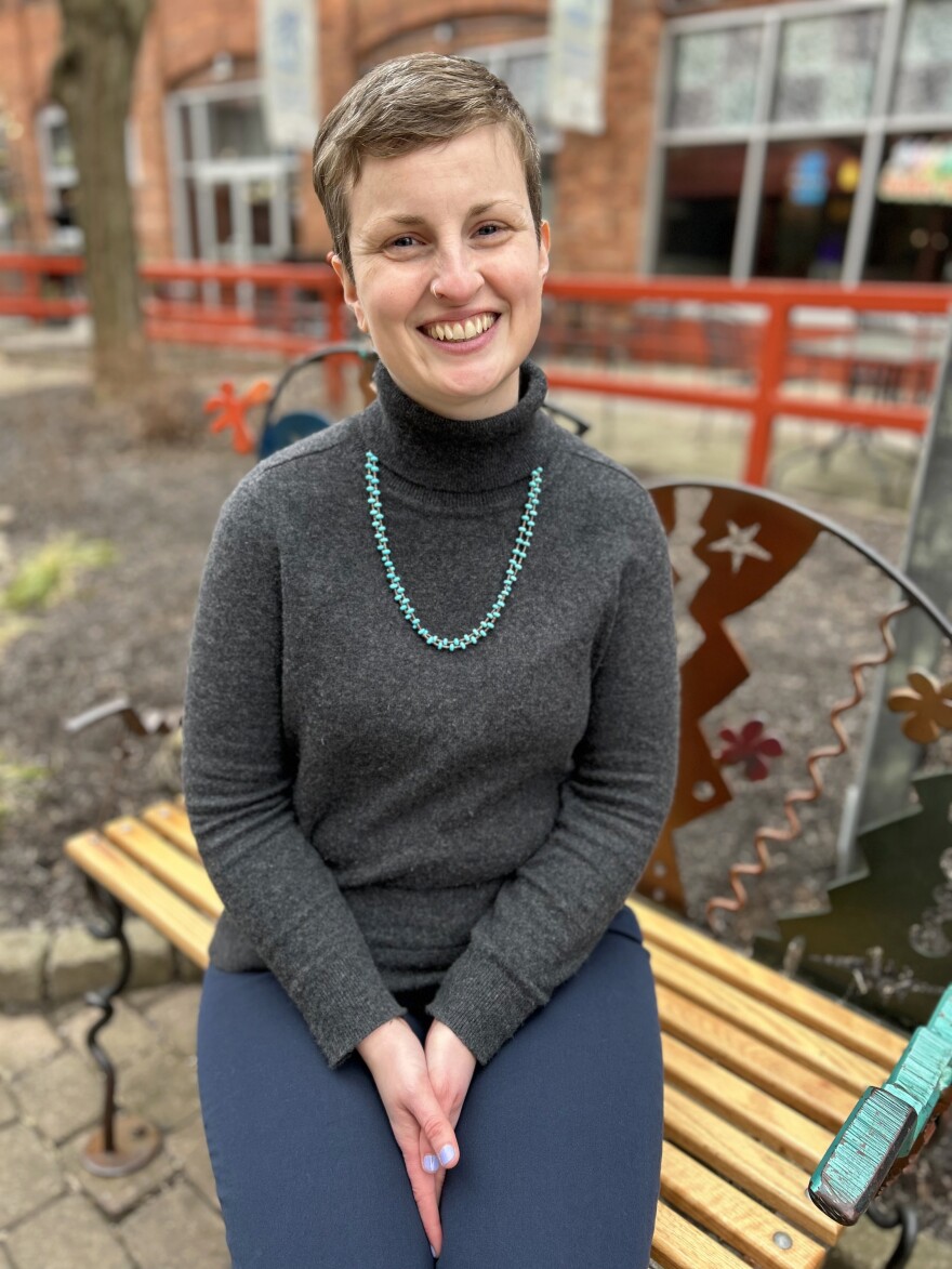 A woman with short light brown hair wearing a necklace of teal colored beads, a grey sweater, and navy blue pants sits on a wooden bench in Village Gate Plaza