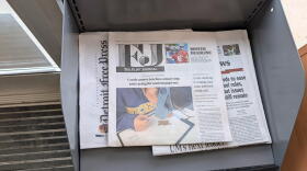 The Flint Journal sits on top of a copy of The Detroit News and the Detroit Free Press in a grey, metal newsstand