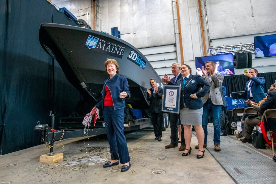 A woman in a blue suit and red shirt stands in front of a boat while holding a broken champagne bottle during a publicity event.