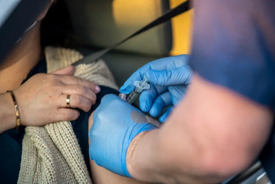 A woman holds her sleeve up as a health worker wearing blue gloves sticks a needle into the woman's upper arm.