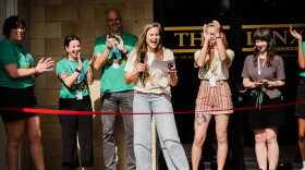 A light-skinned person holding a microphone and standing in front of a ribbon at an opening ceremony. Behind her are a group of people clapping.
