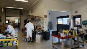 In a large room with white concrete walls and large doors and windows, workers in white suits sort materials. Around them, items of all sorts sit on carts and countertops. 