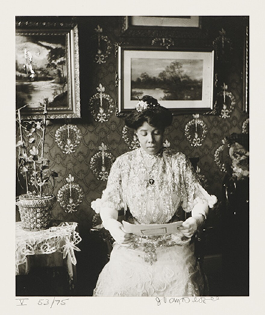 A woman dressed in fine clothing sits in a chair in what looks like a parlor