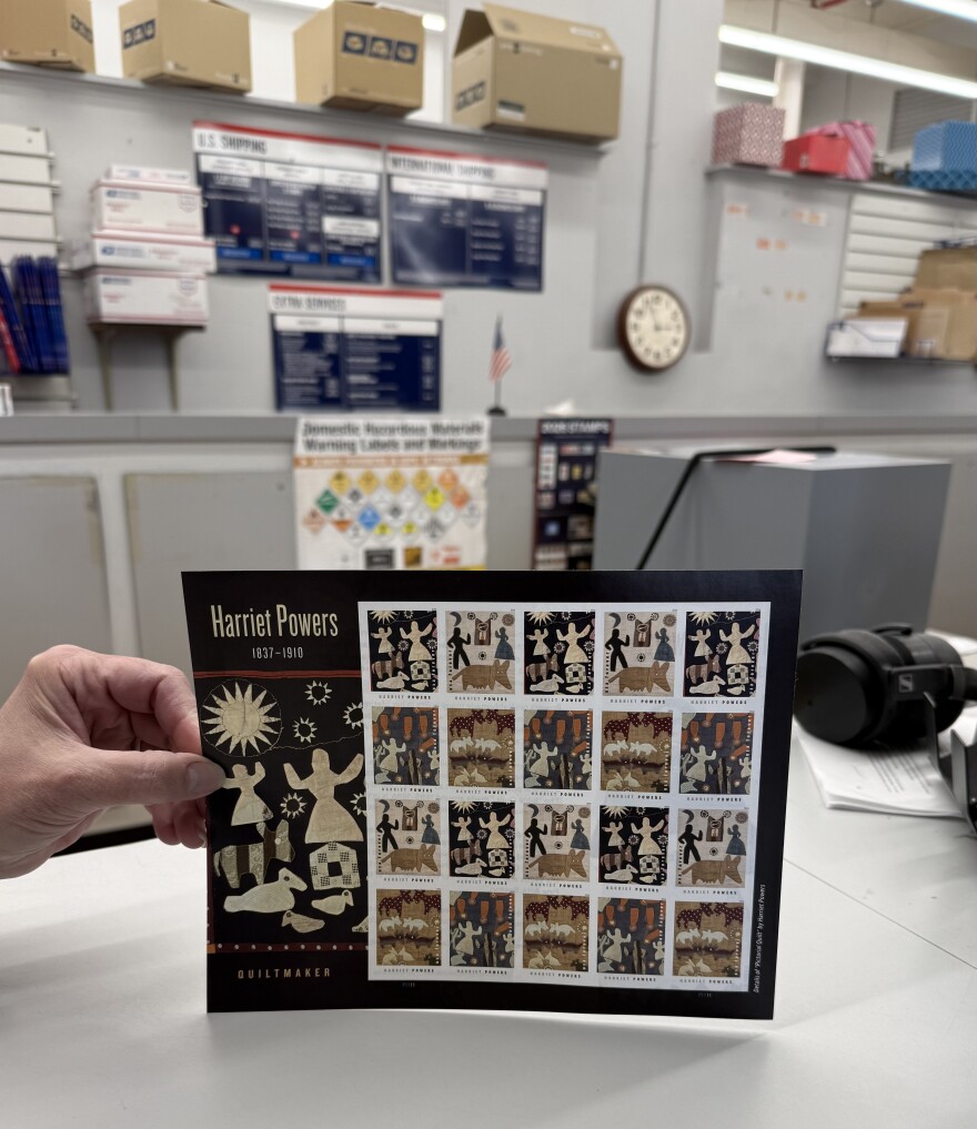 A hand holds a USPS stamp sheet, labeled “Harriet Powers, 1837–1910, quiltmaker,” at a post office counter. Shipping signs, stacked boxes, a wall clock, and a pair of headphones are visible in the softly blurred background.