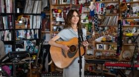 A woman smiles while standing and playing acoustic guitar behind an office desk with shelves of music paraphernalia behind her.
