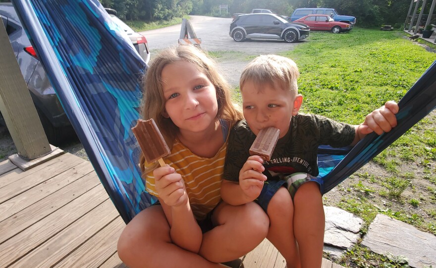 A young girl and boy sitting on a hammock together eating popsicles on a porch in summer