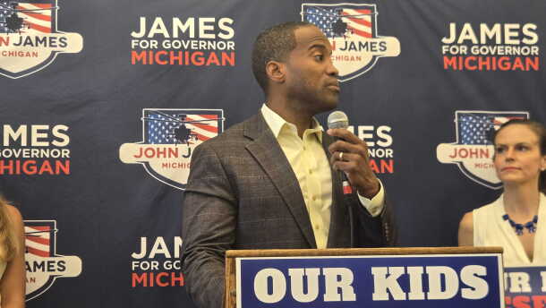 John James stands at a podium, speaking into a handheld microphone. He wears a gray plaid suit jacket and pale yellow shirt. Behind him is a campaign backdrop repeatedly bearing his name and “James for Governor Michigan.” A large sign in front reads, “OUR KIDS DESERVE BETTER.”
