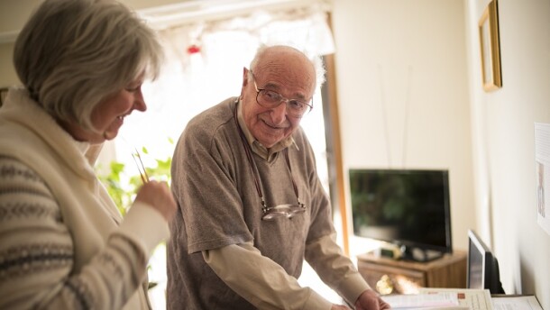 An older gentleman stands in front of a desk and smiles at a woman assisting him. 