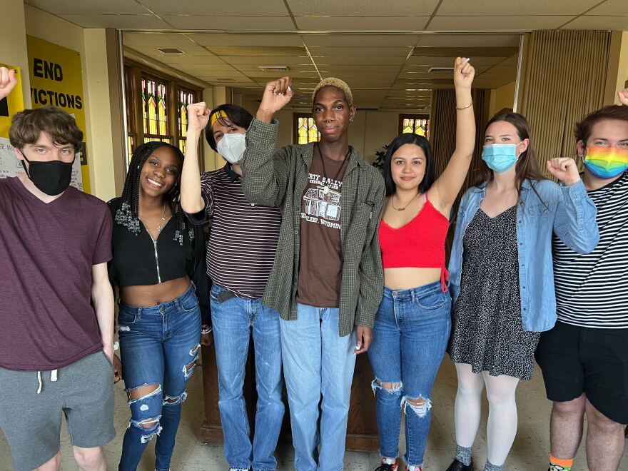 Seven young people stand in a line with their fists raised facing the camera. Half wear masks. 