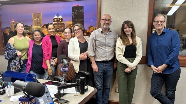 Eight smiling people stand next to each other in a radio talk studio. A print of the Rochester skyline in the evening is behind them. From left to right: a woman with long dark hair, wearing a white headband, a green top, a blue and gray shawl, and jeans; a woman with black curly hair, wearing a black top under a pink blazer, and jeans; a woman with her black hair tied back, wearing a black top under a pink blazers, jeans, and a bracelet; a woman with her dark hair tied back, wearing a brown top; a woman with short gray hair, wearing glasses, and a black top under a white blazer; a bald man with a gray beard and mustache, wearing glasses, a gray button-down shirt, jeans, and brown shoes; a woman with long brown hair, wearing a black top under a white sweater, green pants, and black sandals; a man with very short dark hair, wearing glasses, a dark blue button-down shirt, jeans, and brown shoes