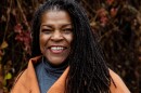 A Black woman wearing an orange overcoat and gray turtleneck in front of an ivy wall.