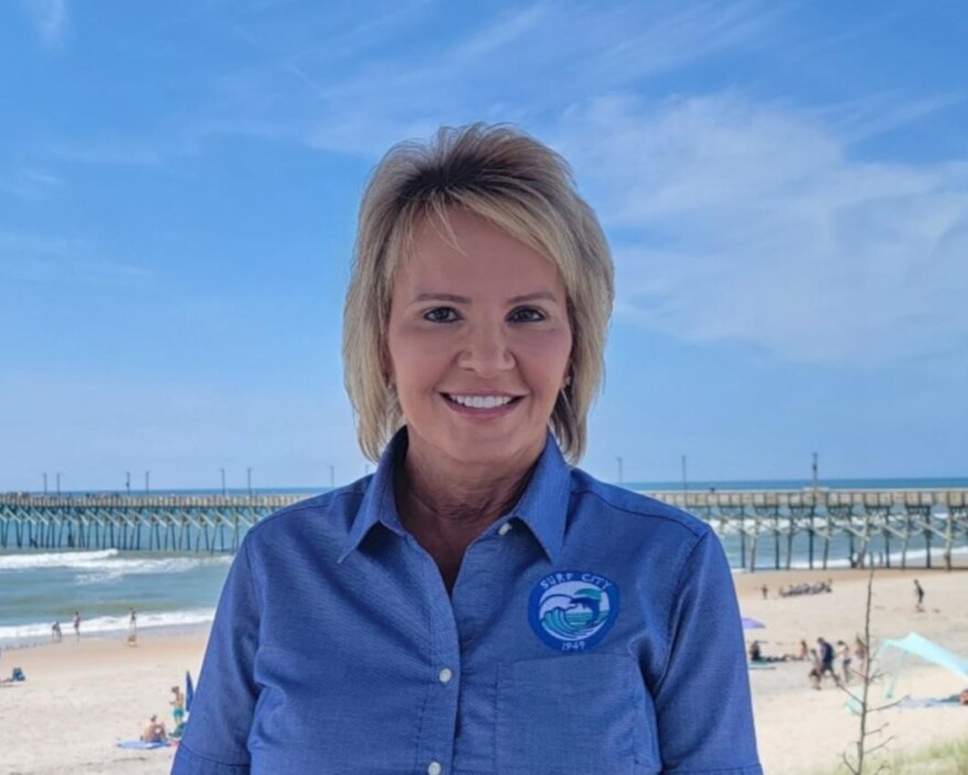 Teresa Batts, a blonde woman in her fifties, poses for a photo on the beach. She's wearing a blue button down with the Surf City Government logo on the right side. Behind her is a fishing pier and golden beach on a sunny summer day. 