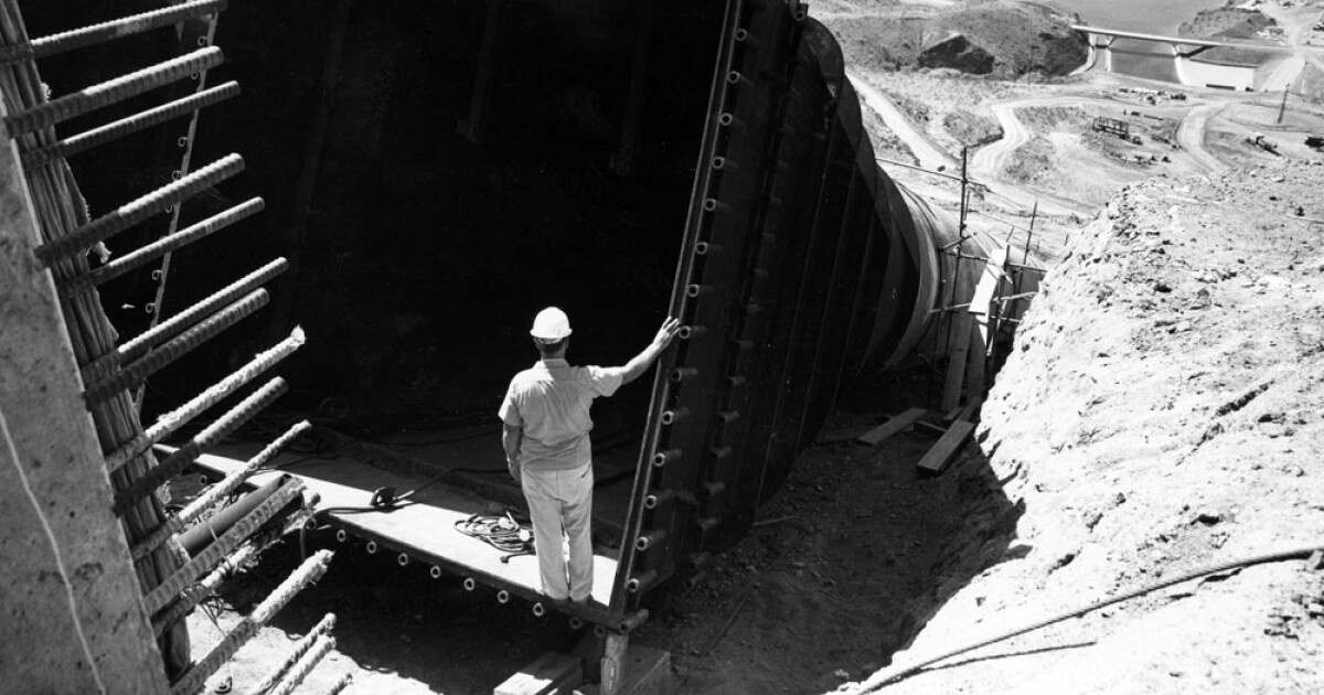 A man in a hard hat stands inside a massive dry pipe, looking down to a reservoir downhill