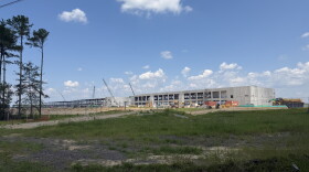 In the center of the photo is a large, concrete building. To the left is the steel skeleton of another giant building. Grass is in the forefront of the picture, and clear skies above the buildings. There are also constructions cranes and other construction equipment. 