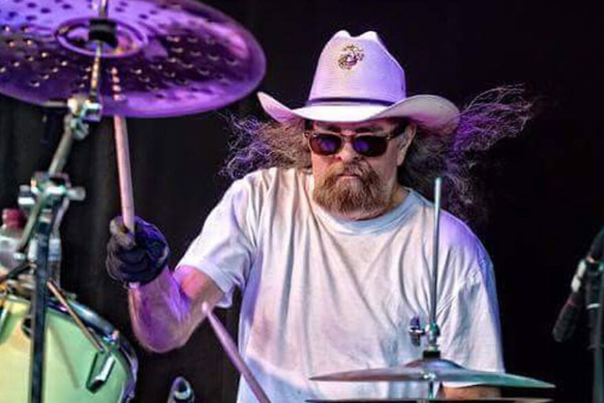 Man with beard and long hair wearing glasses and white tshirt drumming on stage