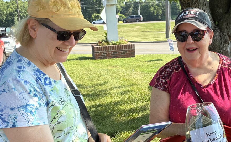 Two women with books and a donation jar at a market.