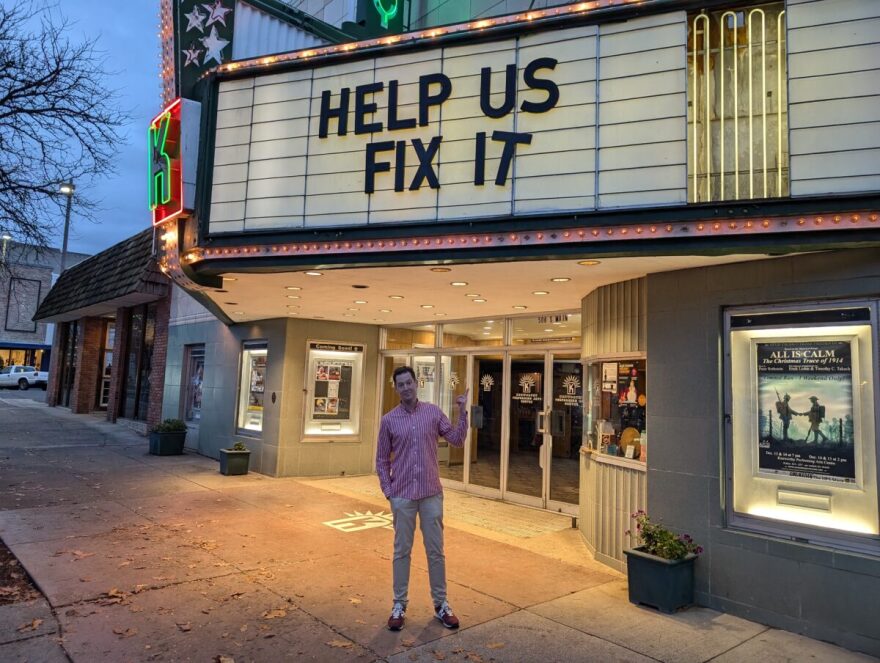 A man in a red and white shirt and khaki pants stands under a large theatre marquee with the words "Help us fix it" written on the marquee.