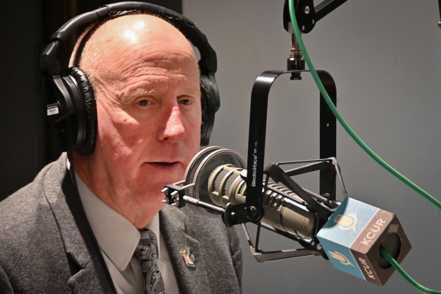 A man wearing a suit and tie sits inside a radio studio talking at a microphone.