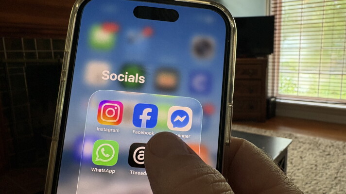 Close-up of a hand holding an iPhone. The screen reads “Socials” and in a collection are icons for Instagram, Facebook, Messenger, WhatsApp and Threads, all apps controlled by Meta.