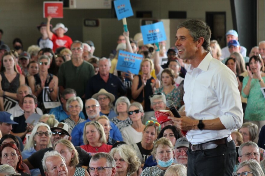 Beto O'Rourke smiles as he stands on stage with an audience behind him, wearing a white button-down shirt and black pants. 