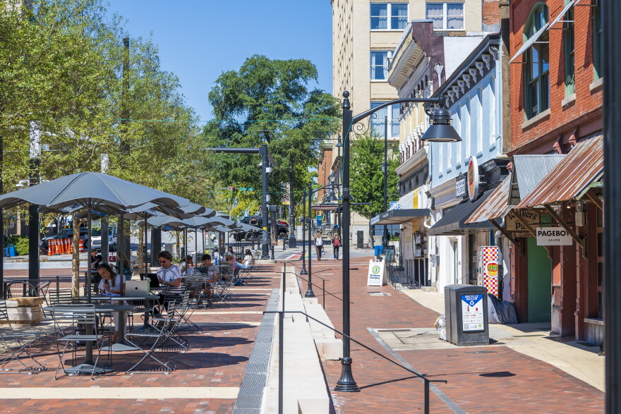 a view down a brick-paved pedestrian space on Aollege Square in Downtown Athens, GA. To the left is a row of umbrella-covered tables. To the right is a row of storefronts. Between them are small flights of three steps down from the tables to the storefronts.