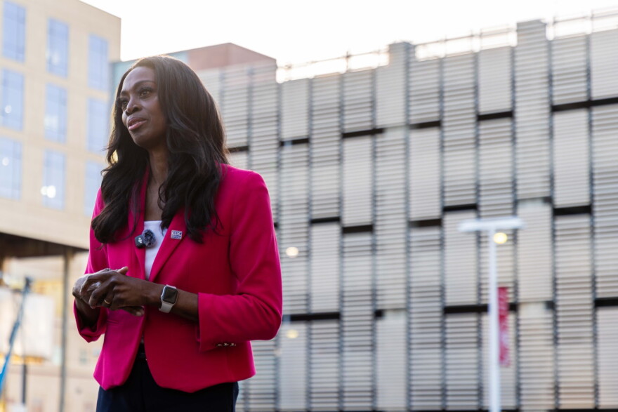 A woman in a pink blazer and a small microphone clipped to her lapel stares ahead.