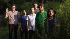 Six people stand in an overgrown field looking at the camera with trees in the background.