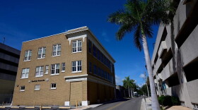 A tan, three-story building stands on the left, next to a road lined with palm trees, a parking garage on the other side of the street.