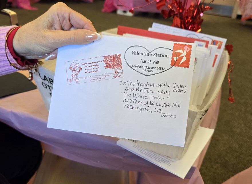 A woman's hand holds a white envelope decorated with red Loveland seal and a heart-shaped Loveland Valentine stamp. The envelope is addressed to the President of the United States and the First Lady.