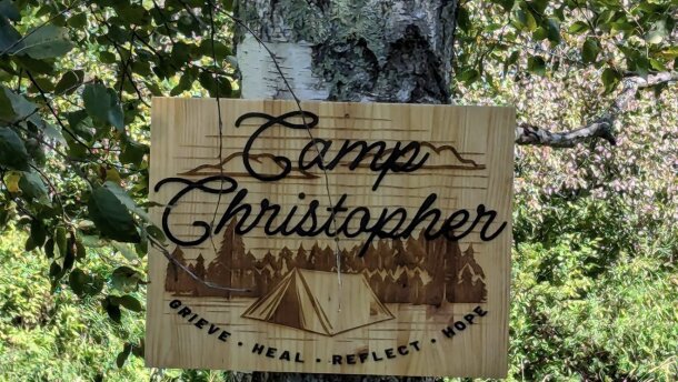 A wooden sign reading "Camp Christopher: Grieve, Heal, Reflect, Hope" is on a birch tree with vegetation in the background. 