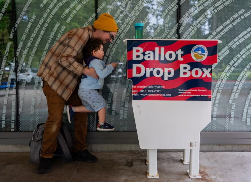FILE - Matt lifts up his three-year-old son, Theo, to cast his ballot into the Hillsboro Public Library-Shute Park ballot drop box in May 2026. Election officials are increasingly recommending voters plan to use drop boxes rather than mailing their ballot.