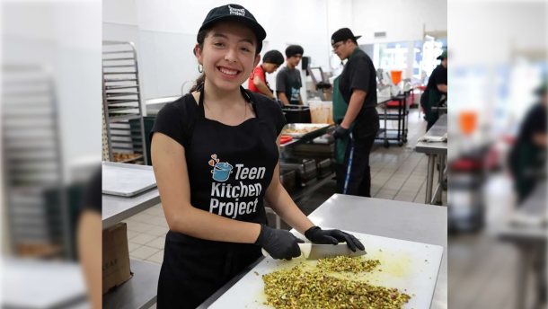 A smiling young woman with a baseball cap and apron wears black food-service gloves as she chops a pile of something on a white cutting board. Behind her more young adults work in a commercial kitchen filled with stainless steel counters and pans. 