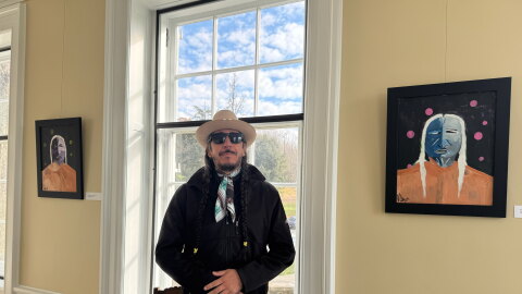A man in a black shirt named Travis Mammedaty (of the Seneca-Cayuga Tribe) stands by his art at the exhibit.